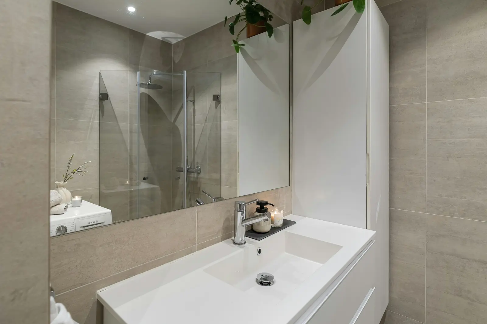 Lavabo blanco frente a un espejo, con revestimiento beige y ducha acristalada al fondo