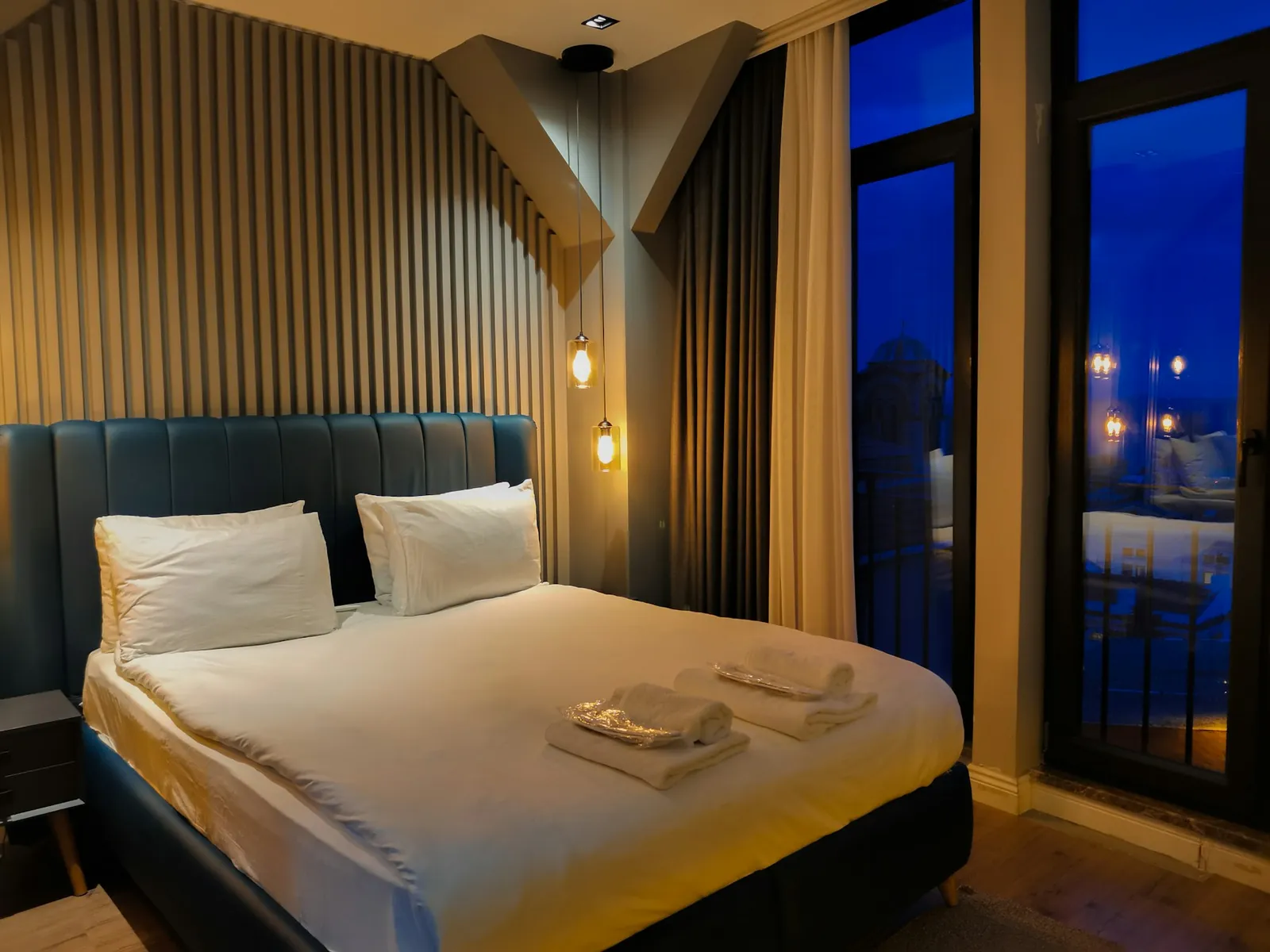Habitación de hotel con cama doble, cabecero tapizado, lámparas colgantes y ventanales