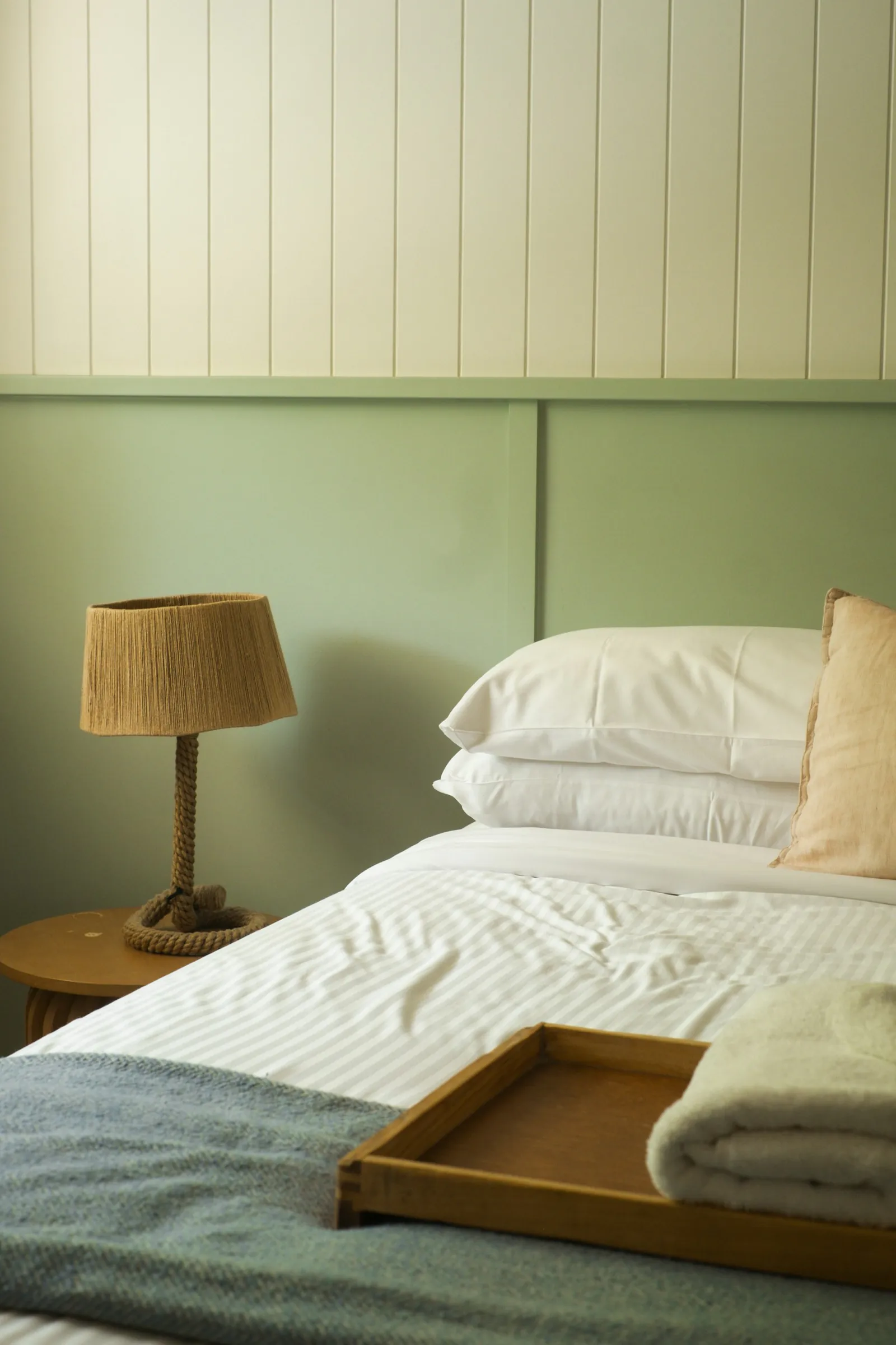 Lámpara de mesilla junto a una cama con cabecero verde y textiles claros